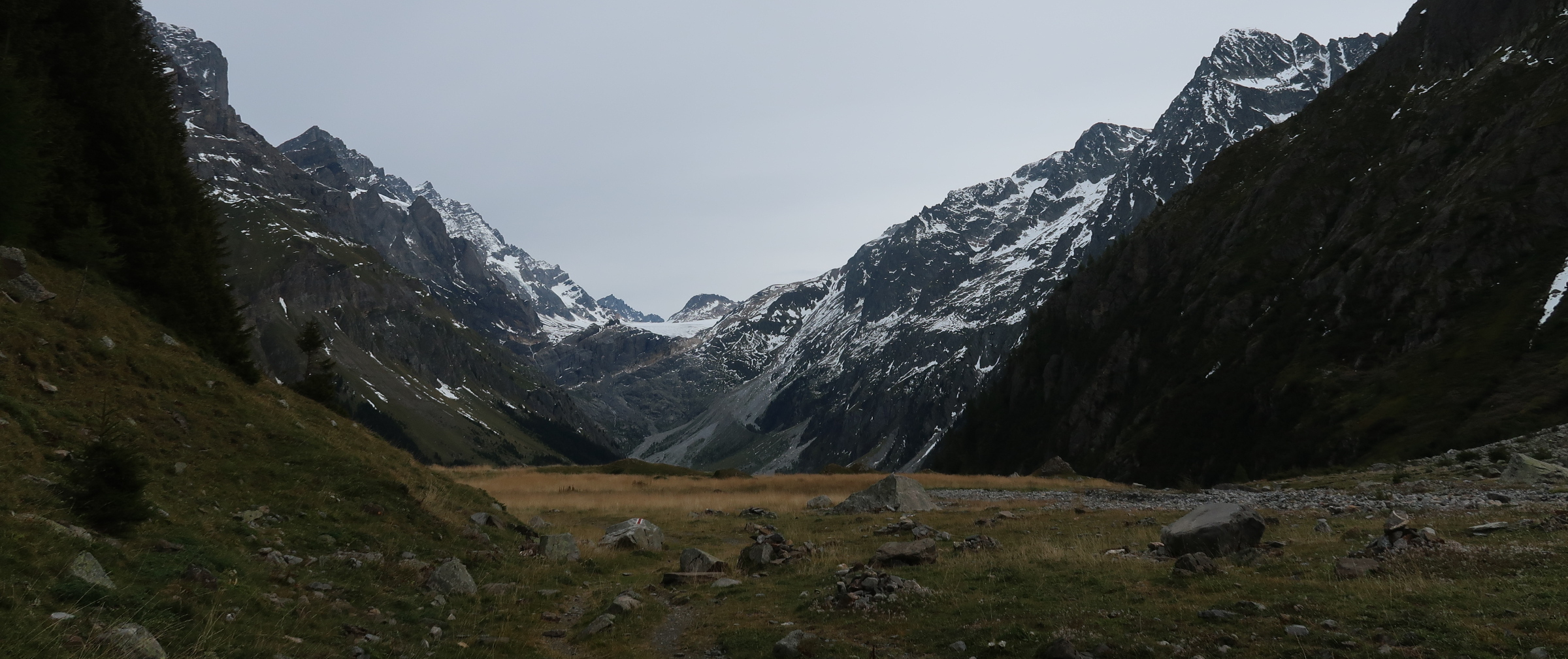 A flat, rock-strewn plain forming a high mountain valley with snowy mountains at one end and steep cliffs on either side